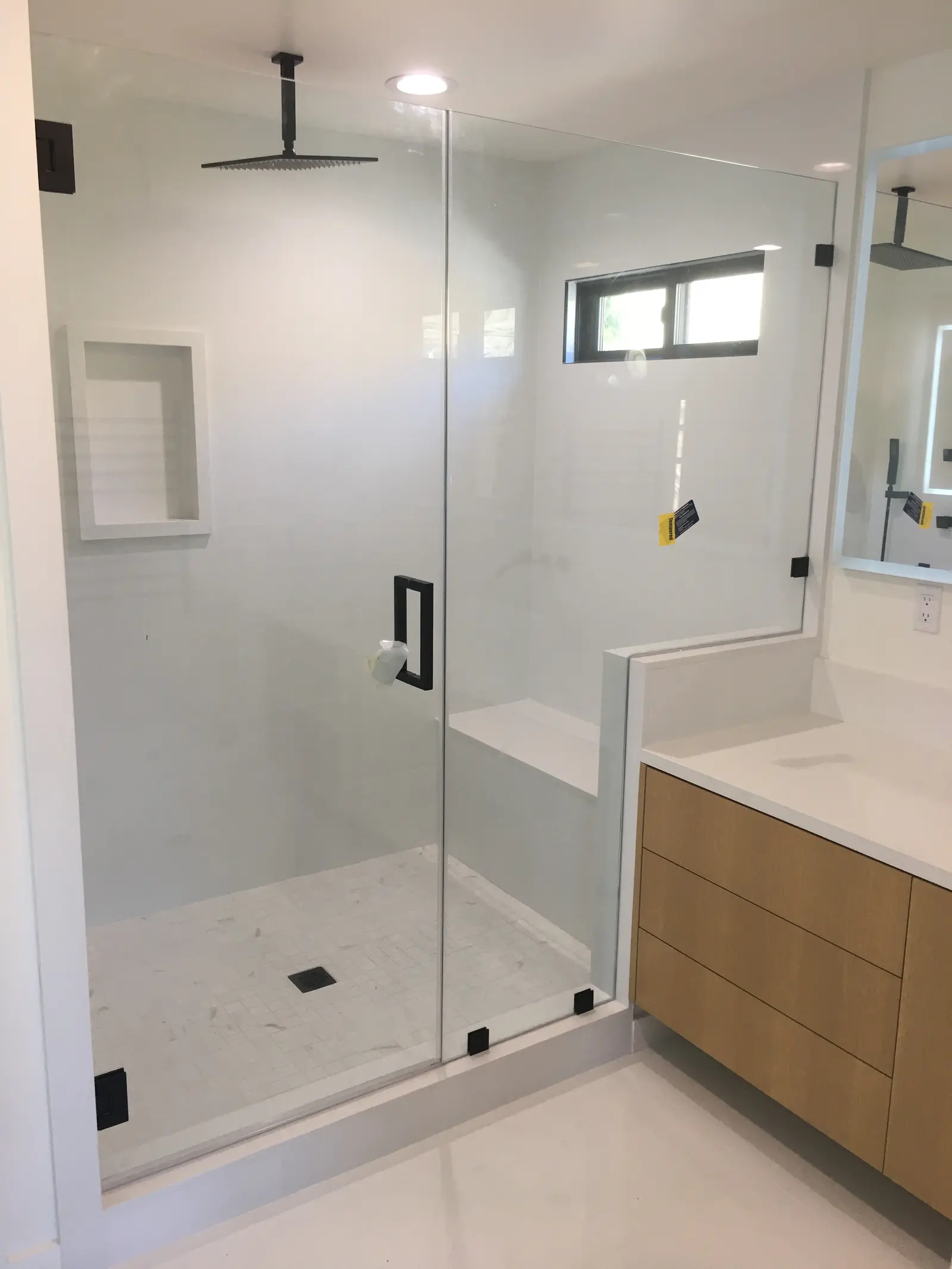 Frameless shower enclosure with matte-black hinges, a corner bench and a white oak vanity