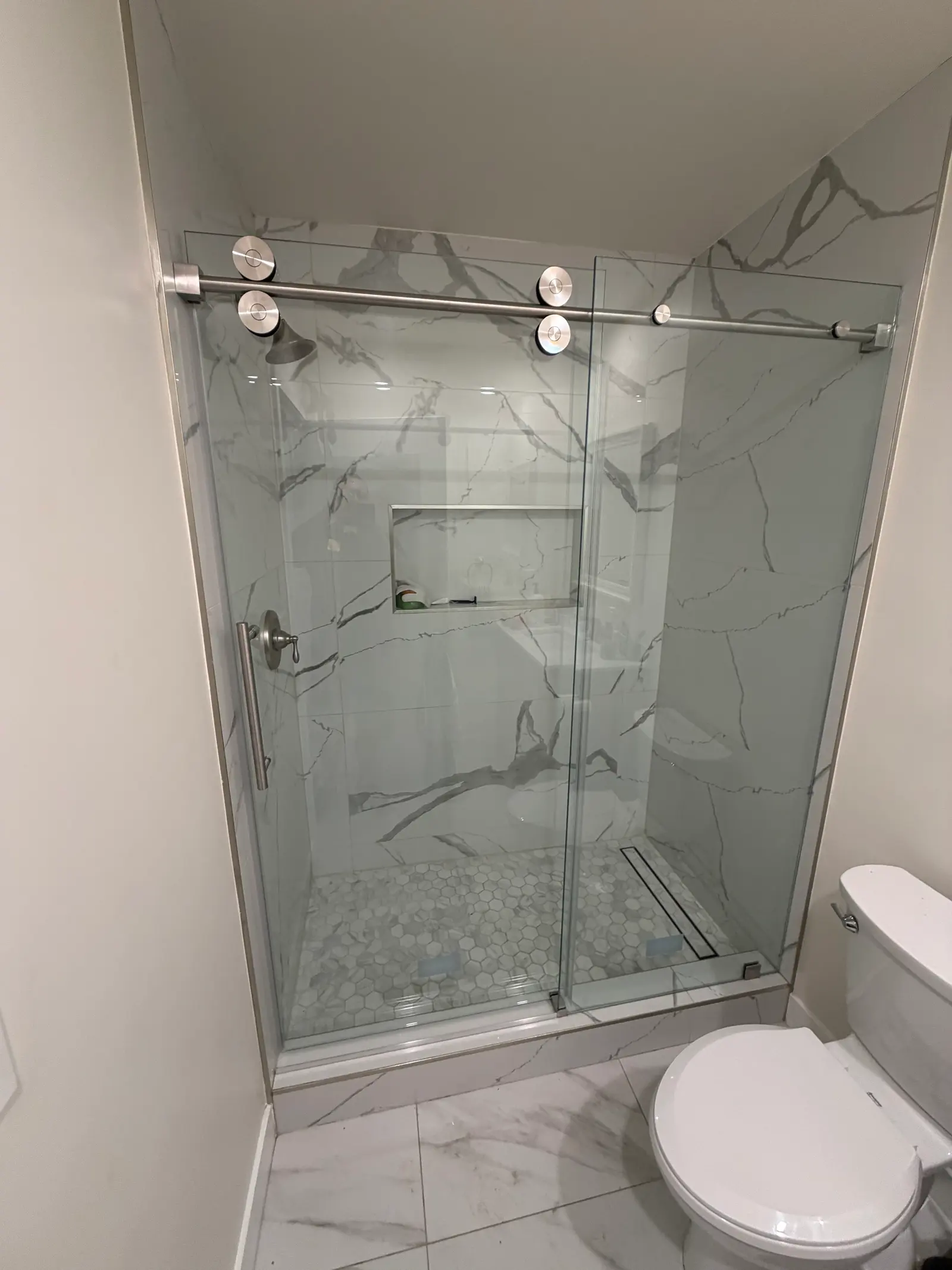 Sliding glass shower door on an exposed brushed-nickel track over a marble-look alcove