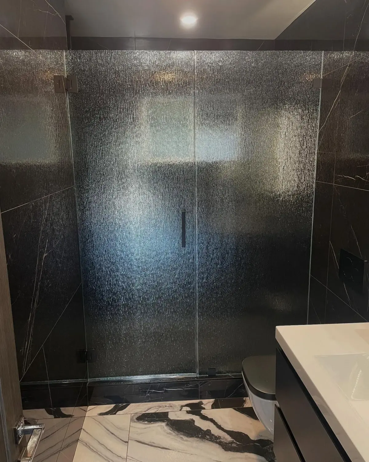 Rain-textured glass shower doors with a slim dark handle against black marble walls